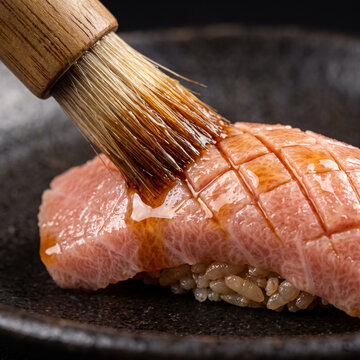 Chef brushing savory nikiri soy sauce onto scored fatty otoro tuna nigiri sushi on black ceramic plate
