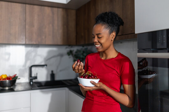 Black woman in red shirt smiling while holding bowl of cherries in modern kitchen