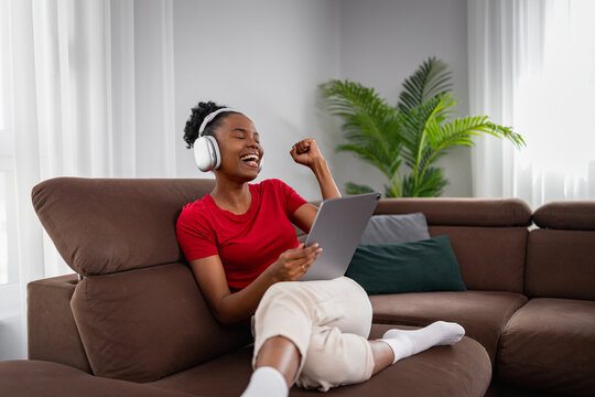 Black woman listening to music on tablet and celebrating while relaxing on sofa