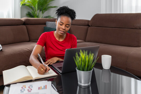 Black woman working on laptop at home taking notes and planning finances on living room table