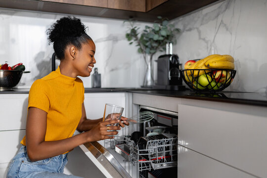 Black woman loading dishwasher while smiling in modern kitchen with fruit bowl and coffee maker