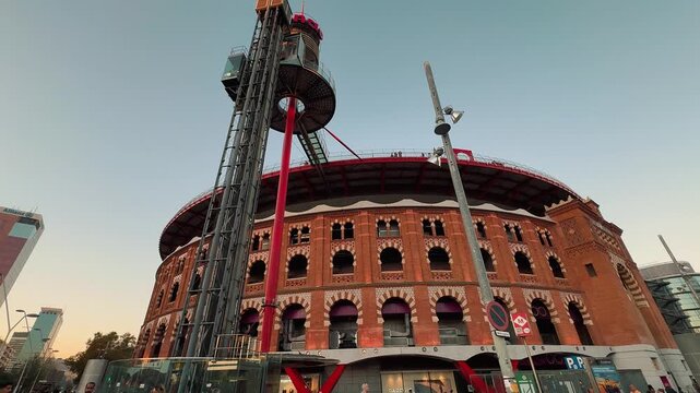 Exterior wide shot of Arenas de Barcelona, a repurposed historic bullring turned shopping center, featuring an iconic outdoor elevator tower and people walking at sunset in Catalonia, Spain.

