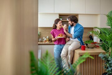 Young couple laughing and sharing croissants while enjoying breakfast in modern kitchen