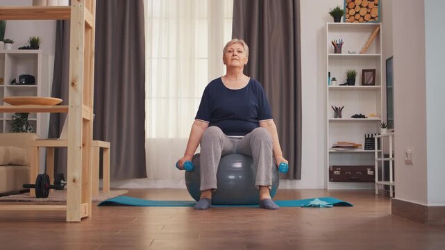 Senior woman performs seated lateral raises with dumbbells on a stability ball at home, combining shoulder strength, balance and active aging.