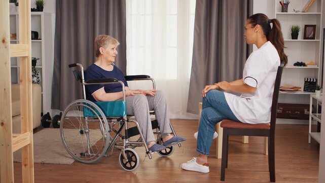 Nurse speaks with a senior woman using a wheelchair at home during a supportive care conversation with attentive listening and personal assistance, with a focus on mobility and home care.