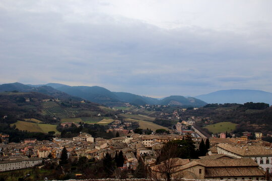 Panoramic viewpoint overlooking Spoleto, with human settlements and plenty of houses and buildings in the foreground of the picture, opening to thickets, hills, distant mountains, under a cloudy sky