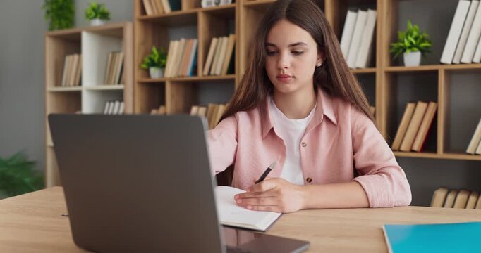 Student teenager girl writing while studying laptop. At a desk in the library, she takes notes from laptop, books nearby and homework and learning. Education and personal productivity concept.
