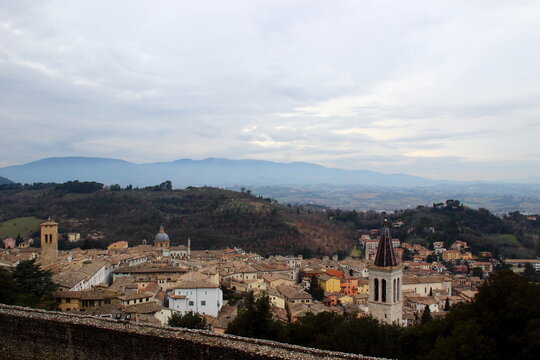 Scenic view of Spoleto with terracotta rooftops and church towers in front of thickets, hills, distant mountains, creating a particular, longing atmosphere, under a cloudy, airy, spiritual sky
