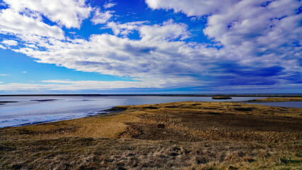 A vast coastal lagoon and golden tundra flats stretch toward the distant horizon under an expansive, cloud-filled blue sky in Iceland © Gregory