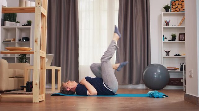 Senior woman lies on a yoga mat and raises her legs during a home core exercise focused on abdominal strength, control, mobility and active aging to support healthy aging and senior fitness.