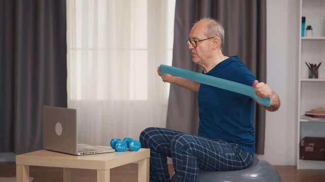 Senior man pulls a resistance band across his chest while watching an online workout on a laptop, showing upper-body exercise and active aging at home to support mobility and senior fitness.