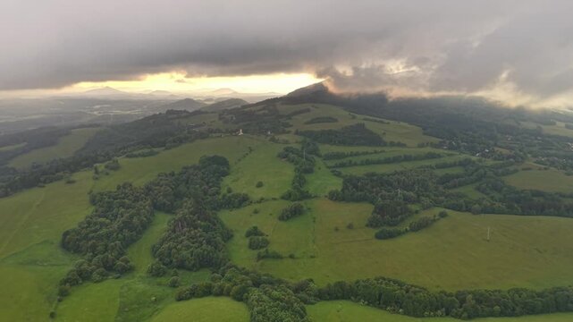 Panoramic aerial view of patchwork farmland across rolling hills, with tree clusters, green pasture, distant village, and dramatic sunset clouds creating golden glow over ridges and valleys