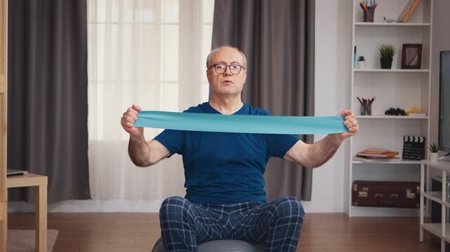 Senior man pulls a resistance band at chest level while seated on a stability ball, combining upper-body strength, balance and active aging at home.
