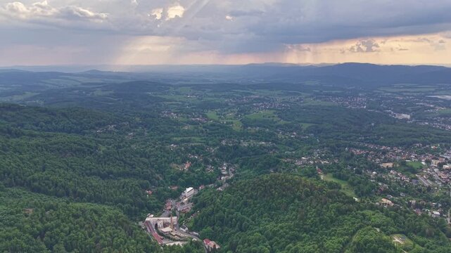 Serene rural aerial panorama showing patchwork farmland, dense forests on rolling hills, and scattered towns as sunbeams break through cloud cover over distant mountain ridges