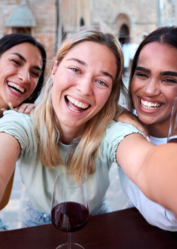 Vertical. Three cheerful multiracial female friends are smiling and taking a selfie while drinking glasses of red wine at a restaurant in a charming european town