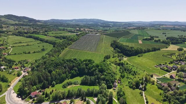 Bright aerial view of patchwork farmland across rolling countryside, with neatly arranged fields, forested areas, and small villages connected by winding roads and paths in clear daylight