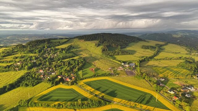 Birds eye view of broad rural landscape showing quilted fields in yellow and green, gentle hills, and small village nestled among trees, with aerial, landscape, field, farmland detail