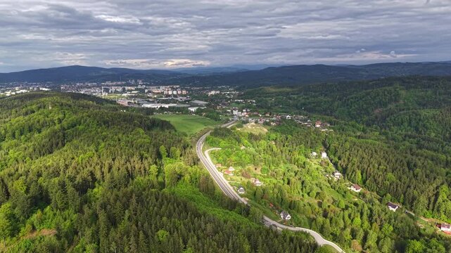 Overhead view shows dense evergreen forest on rolling hills, with winding road threading through green valleys toward compact town, with forest, hill, town, road detail, showing landscape, aerial