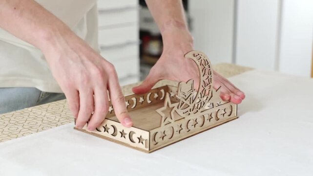 A young, unrecognizable man, standing at a table, presses the side pieces into the holes in the bottom with both hands, assembling a carved wooden tray. Close-up, side view with slow motion. Concept: 