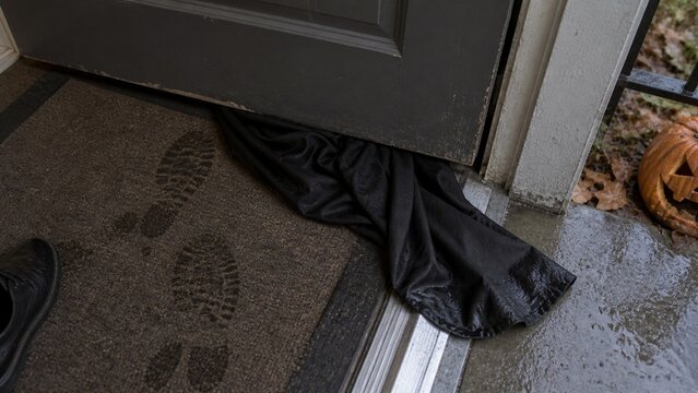 Black fabric extends from under a closed grey door onto a brown doormat, while a carved orange pumpkin sits on a wet concrete porch