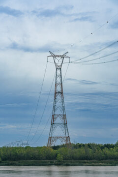 Tall high-voltage power line pylon standing near a river shore against a cloudy sky, distributing electrical energy through overhead cables across a scenic natural landscape