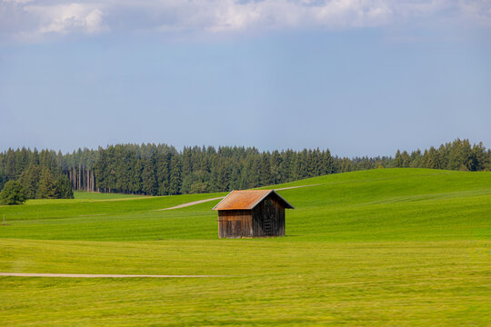 Bavarian countryside landscape, A traditional wooden barn standing alone on a rolling hill, Lush green grass meadow against majestic Alpine mountains under blue sky and white clouds, Bavaria, Germany.