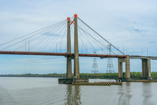 Wide side view of a large cable-stayed suspension bridge spanning over a calm river, featuring tall concrete towers, red roadway deck, and electric power line pylons nearby