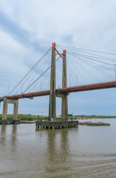 Vertical low-angle view of a cable-stayed bridge featuring concrete support pillars, red deck, and steel suspension cables spanning over a wide river against a cloudy sky