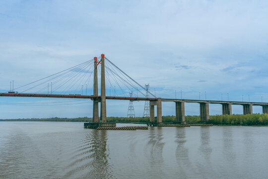 Distant wide landscape shot showing a long cable-stayed bridge spanning across a broad river with overcast skies and power pylons visible in the background along the green shore