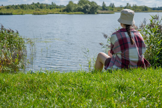 Woman in a light sun hat and burgundy checked shirt sits on green grass beside a river framed by dense reeds on both sides. The secluded setting represents outdoor relaxation, solitude, nature escape