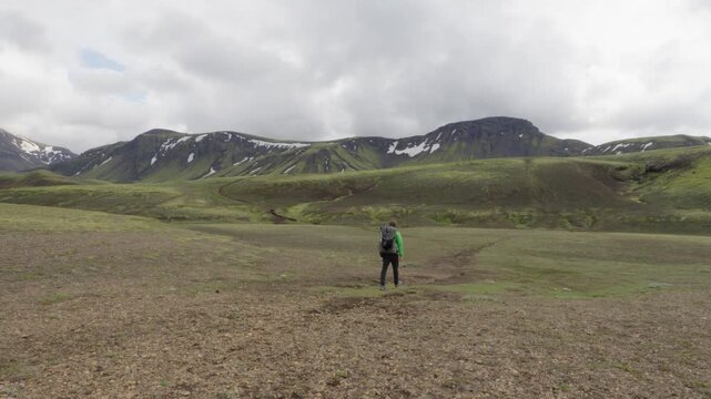 A lone hiker walks on the famous Laugavegur trail on Iceland, traversing a remote and vast mountain landscape with lush green hills and snow patches, showcasing adventure and solitude.
