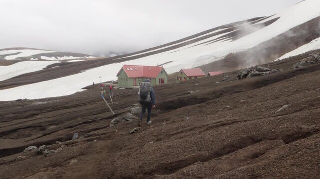 Hiking on the famous Laugavegur trail in the rugged, volcanic landscape of Iceland. A solitary adventurer walks towards Hrafntinnusker mountain refuge huts amidst snowfields and geothermal steam.