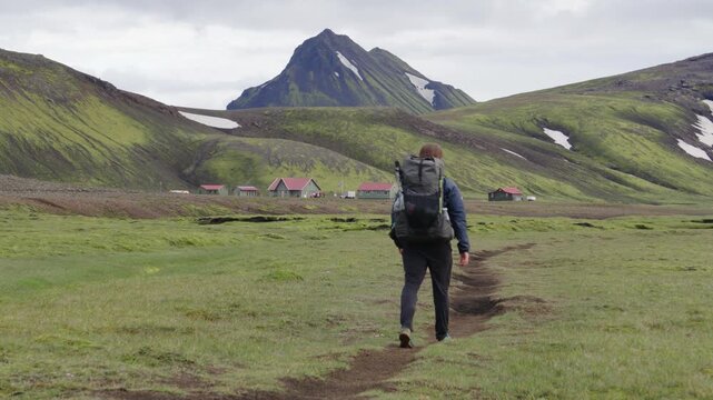 A lone hiker treks along the iconic Laugavegur trail on Iceland, surrounded by vast green, rugged mountains, patches of snow, and &Aacute;lftavatn camp site. Adventure and exploration in stunning nature.