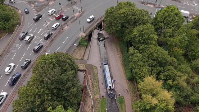 Aerial view of a narrowboat approaching a canal lock in England, where the water level is lowered to allow the vessel to continue beneath a busy road bridge.