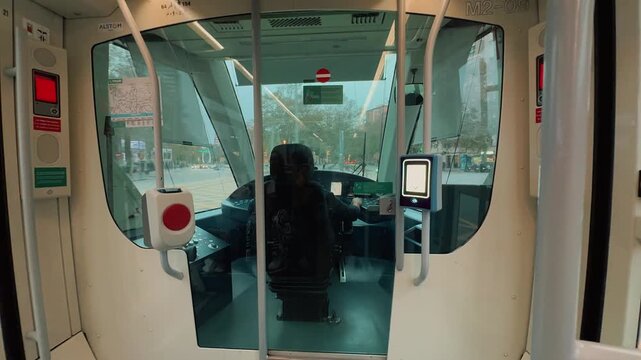 Interior view of a driver operating a modern electric city tram through urban streets, filmed from behind the transparent glass cabin partition inside the passenger compartment during daytime.

