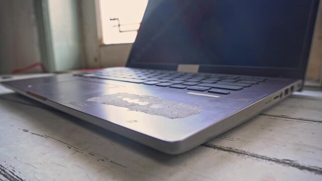 Close up shot of an old worn laptop computer with peeled palm rest on a rustic table by a window, showing hardware wear and tear from long term daily office use.