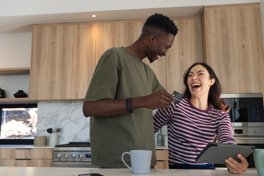 Diverse couple leaning at kitchen island, man holding credit card, woman holding tablet laughing