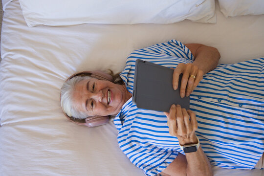 Senior woman lying on bed in striped shirt holding tablet in folio listening with pink headphones
