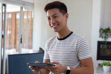 Obraz premium Mid-adult Asian man standing holding tablet with smartwatch in living room monitor showing code