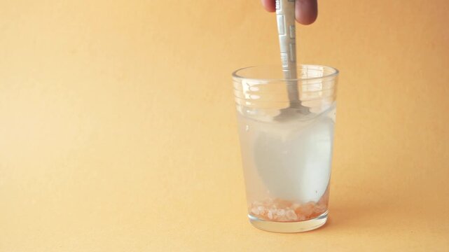 A close up video of a spoon stirring himalayan pink salt in a glass of water on a light brown background