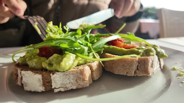Person cutting into fresh avocado toast with arugula and tomato on rustic bread during breakfast outdoors