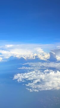 Aerial view of fluffy white clouds and blue sky captured from an airplane window during flight over a landscape with visible land. Slow motion vertical footage, 4K, copyspace