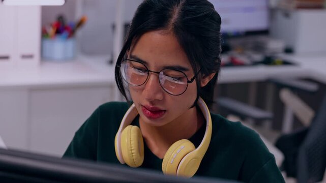 Asian woman wearing glasses and headphones computer workstation, focused office worker and employee engaged with technology, adult person studying or working modern indoor workspace with thoughtful