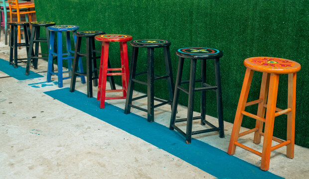 Bright, colorful wooden bar stools stand in a row against a green artificial-grass wall. Street cafe seating area and local pub recreation.