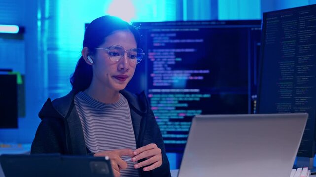 Woman programmer using laptop computer blue lit technology workspace, coding software development screen, focused Asian developer expressing concentration during programming tasks modern office with