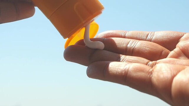 Person squeezing sunscreen onto palm of hand with blue sky in background