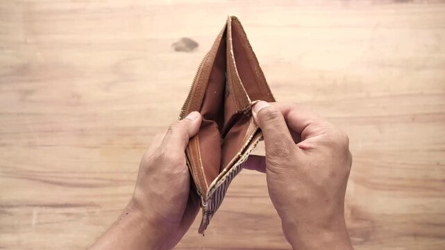 A pair of hands is shown from above, holding an empty brown wallet on a wooden table, emphasizing financial difficulties and lack of money