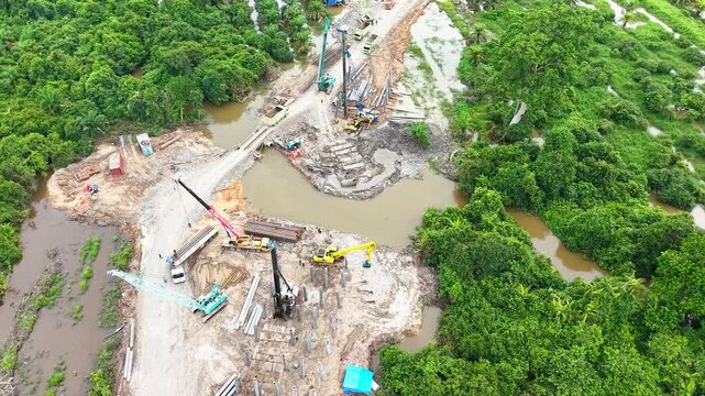 High-angle aerial orbit drone view of bridge construction site over river showing pile driver crane, excavator, heavy equipment, and civil engineering infrastructure development.