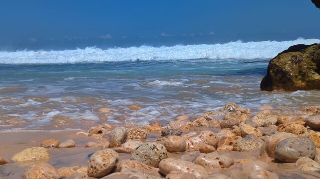 Ocean waves crash against the pebbly shoreline of a tropical region, blending the colors of the sea and the surf into a truly soothing sight.