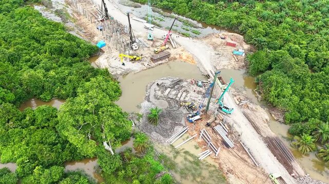 High-angle aerial orbit drone view of bridge construction site over river showing pile driver cranes, excavators, heavy equipment, and civil engineering infrastructure development.